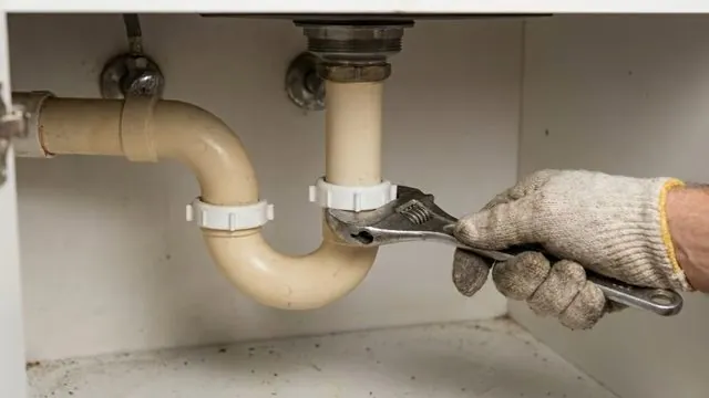 Gloved hand using an adjustable wrench on a beige P-trap under a sink cabinet