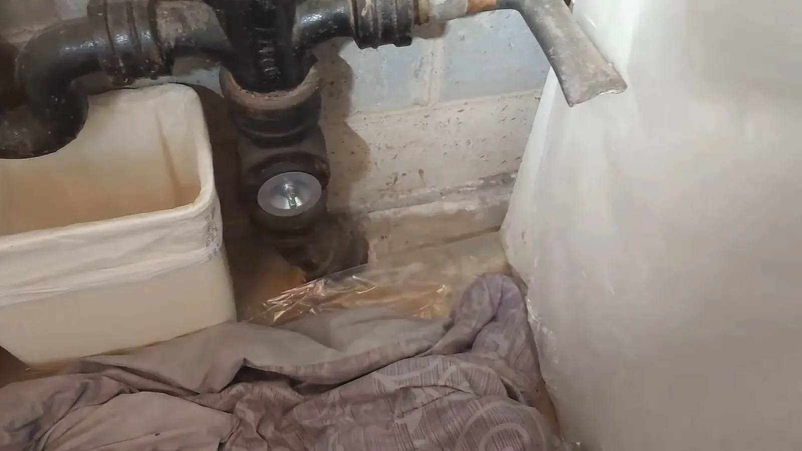 Cast iron drain stack meeting the floor beside a utility sink with a stained cloth on the ground