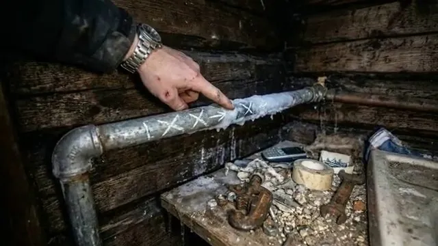 Hand pointing at chalk X marks on a galvanized pipe in a dark crawlspace