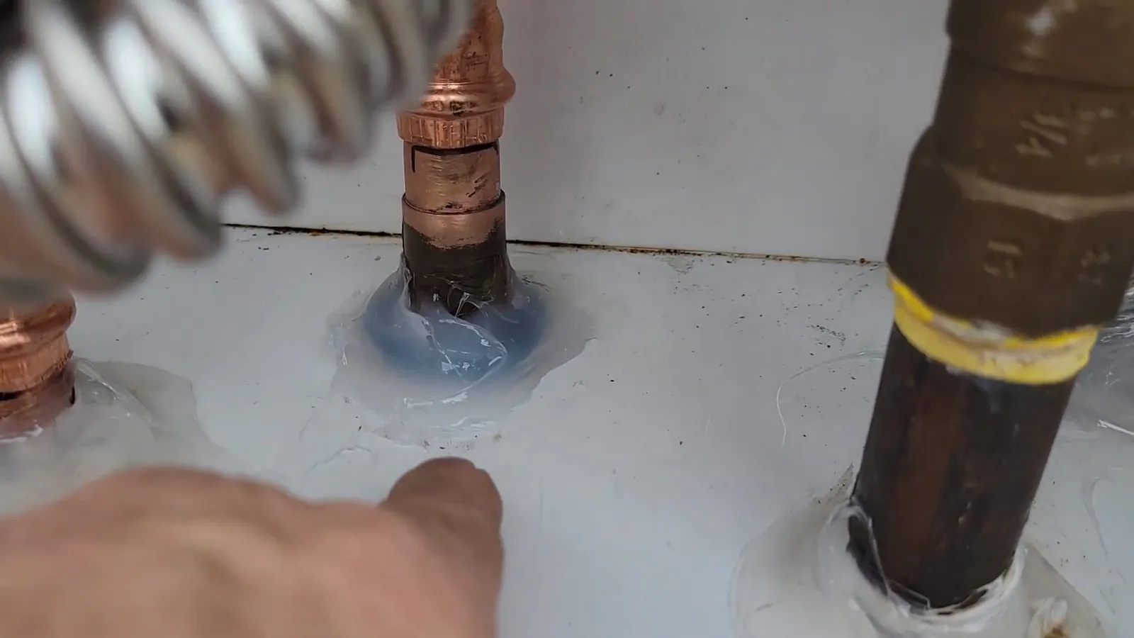 Hand pointing at a copper fitting sealed with blue compound beside a flexible metal line