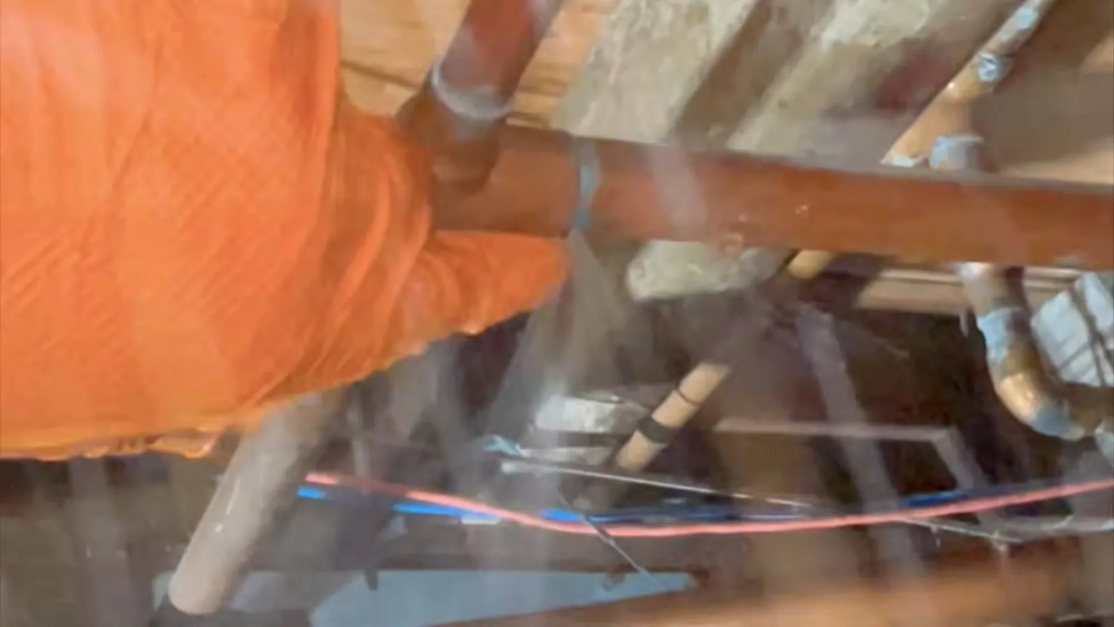 Orange-gloved hand on copper pipes inside a wall with water spraying from the pipe