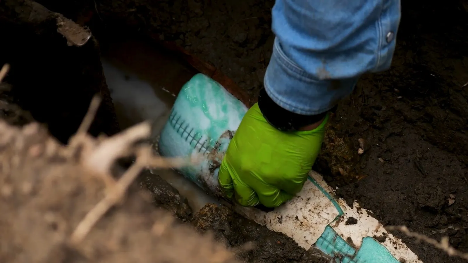 Green-gloved hand on a striped liner pipe lying in a dirt trench