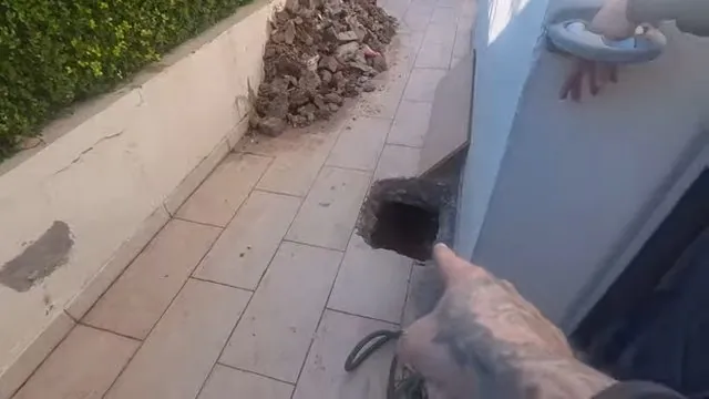 Hand pointing into an open sewer cleanout beside a house wall and tiled walkway