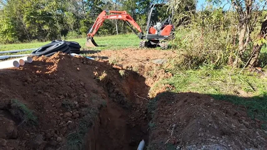 Long dirt trench with a white pipe at the bottom and a small orange excavator behind it