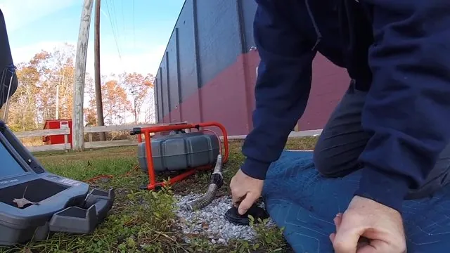 Man kneeling at a sewer camera reel and case set up on grass beside a cleanout