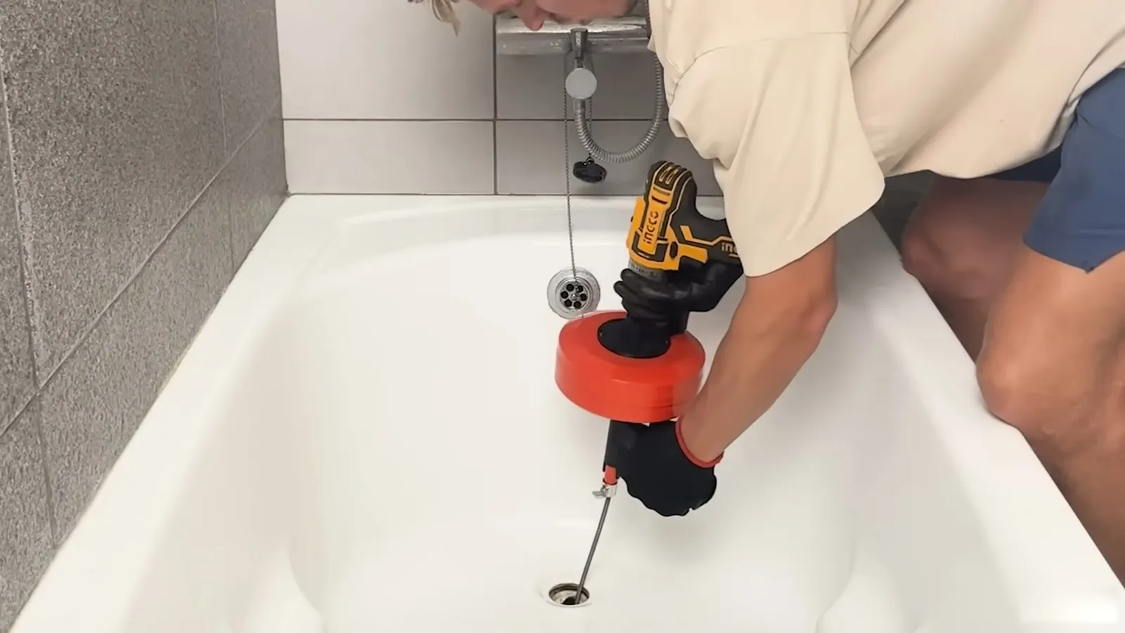 Man using a drill-driven drain auger with a red head in a white bathtub drain