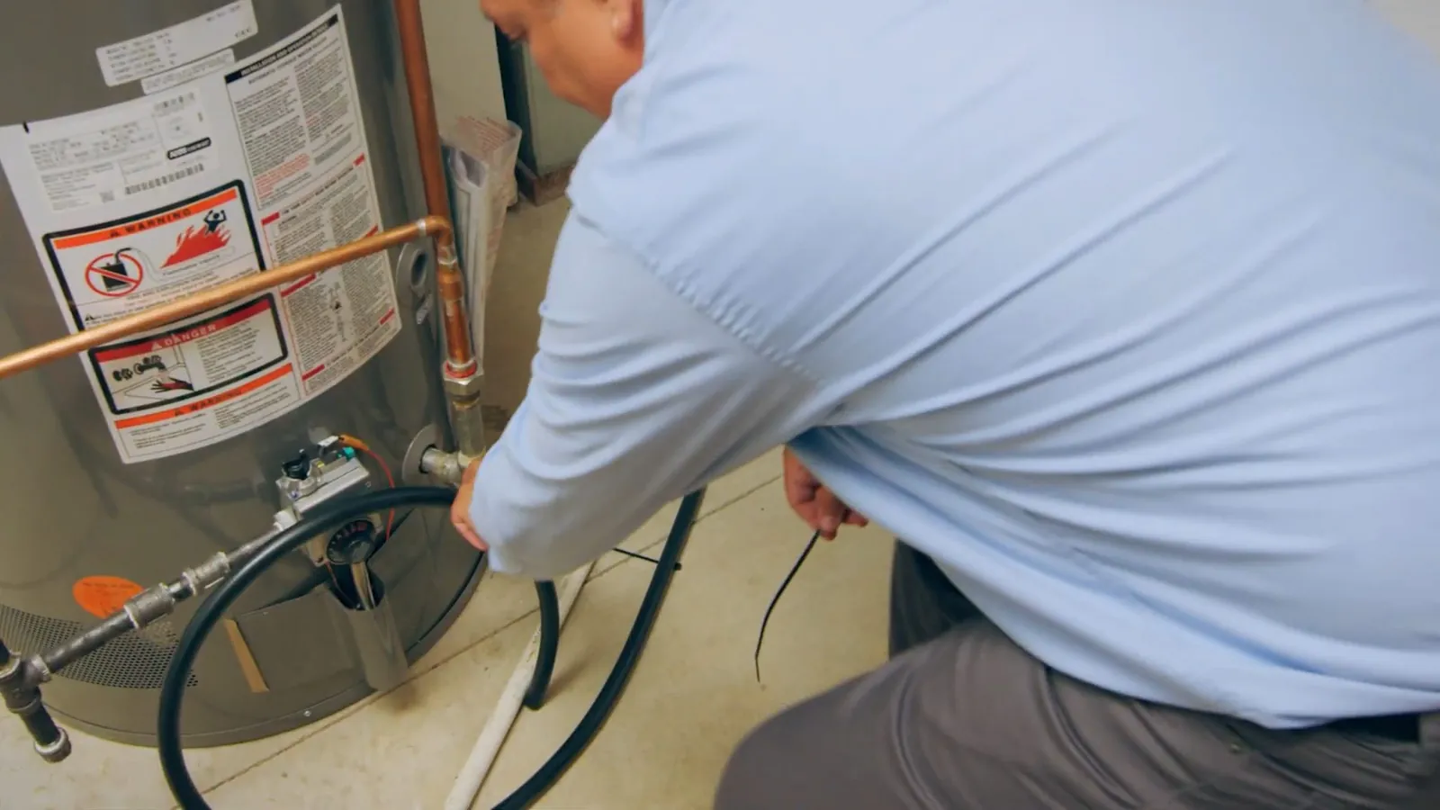 Man in a light blue shirt kneeling at a gas water heater with its connections exposed