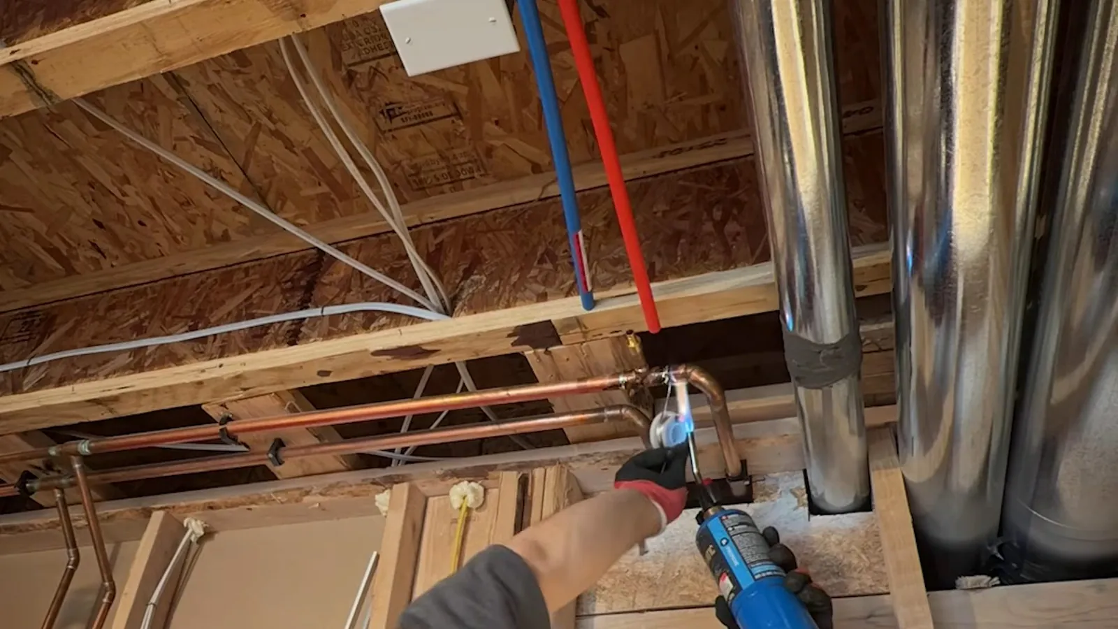 Open ceiling joists with blue and red PEX tubing and copper pipe, and a hand holding a tool