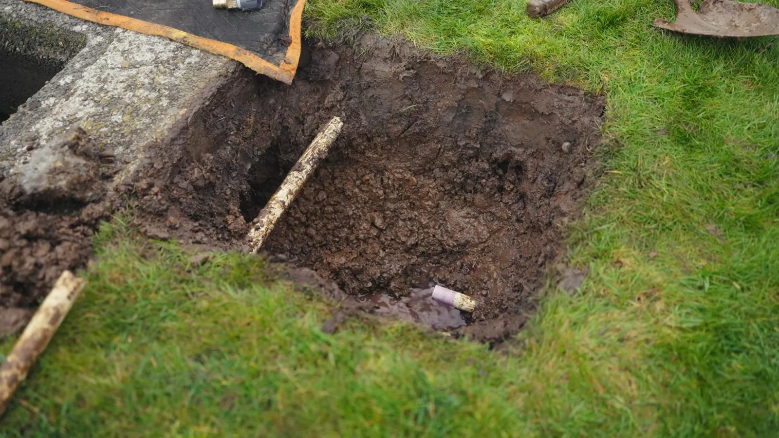 Square excavated pit in a lawn with dark soil and a wooden stick across it