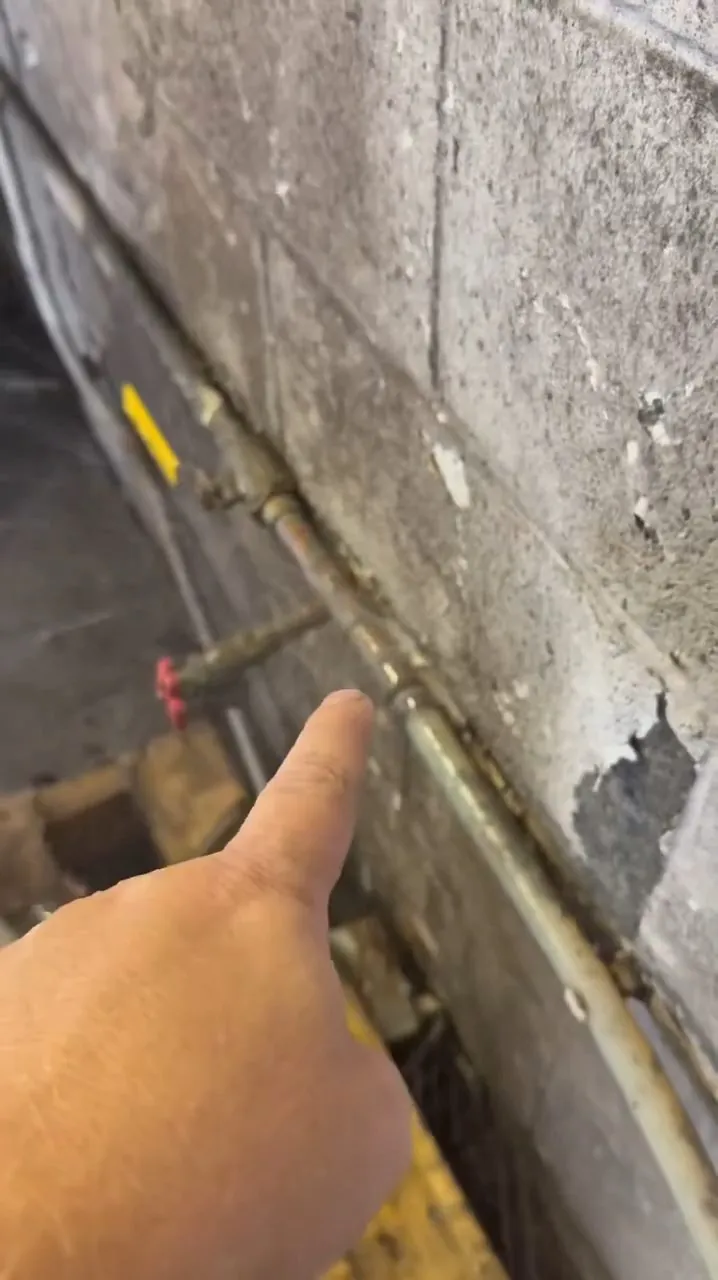 Hand pointing at a pipe running along a stained, peeling concrete block wall