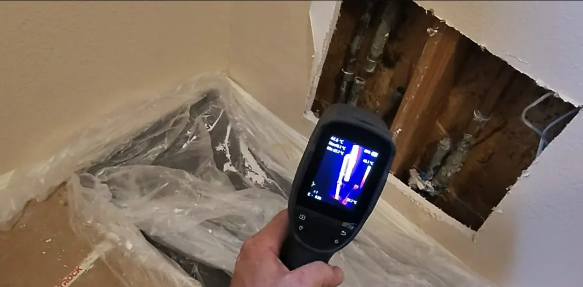Hand holding a thermal camera aimed at an opened section of wall that exposes pipes