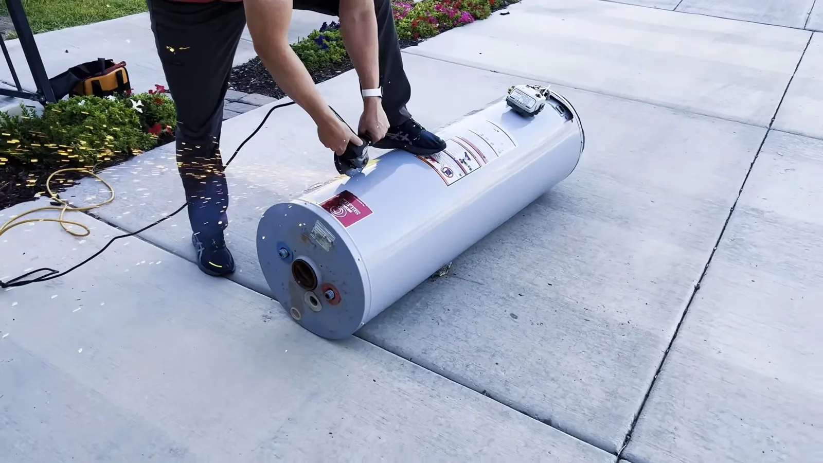Two people tilting a white water heater tank on a concrete driveway
