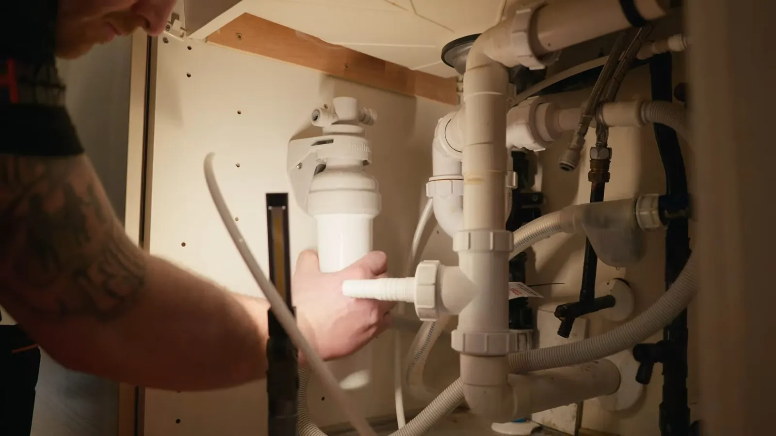 Man holding a garbage disposal under a sink with white drain pipes around it