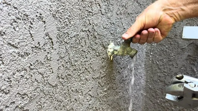 Hand opening an outdoor hose bib on a stucco wall with water running down