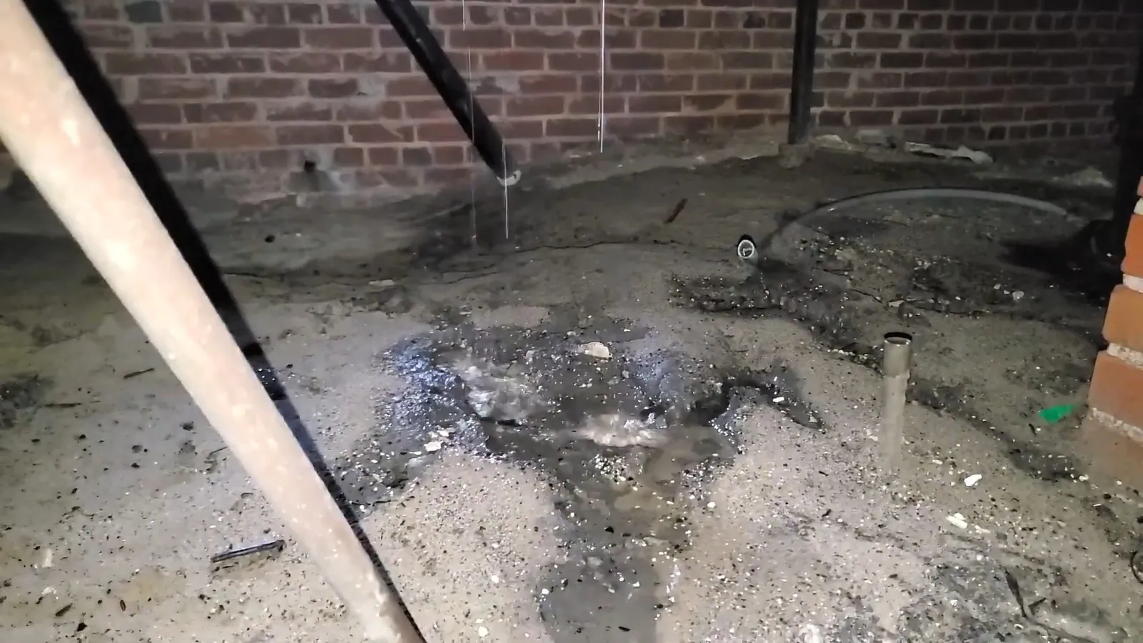 Wet dirt floor with wet patches, pipes and a brick wall in a basement