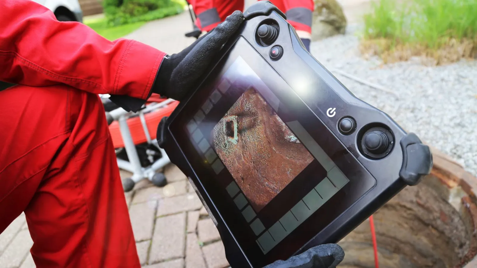 Worker in a red jacket holding a sewer camera monitor beside a yard cleanout